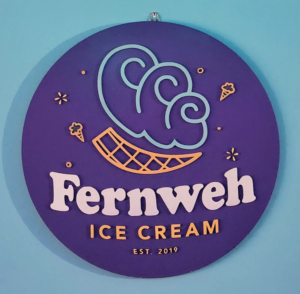 Round purple Fernweh Ice Cream sign hung on a sky-blue wall, with the cone-and-scoops logo mark above the wordmark