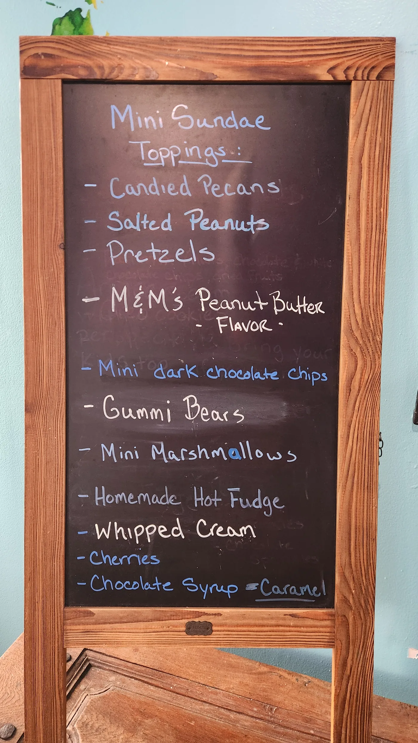Hand-lettered chalkboard listing mini sundae toppings: candied pecans, salted peanuts, pretzels, gummi bears, homemade hot fudge, whipped cream and more